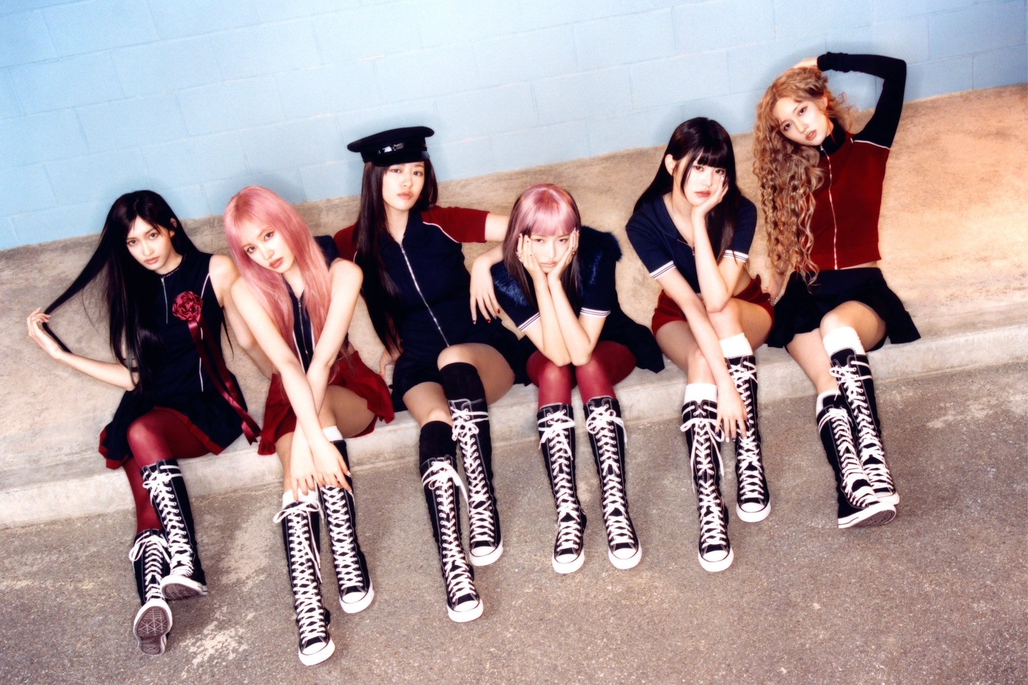 Seis jovens sentadas lado a lado em uma calçada, usando botas pretas de cano alto com cadarços brancos e roupas em tons de preto e vermelho, posando de forma estilosa.