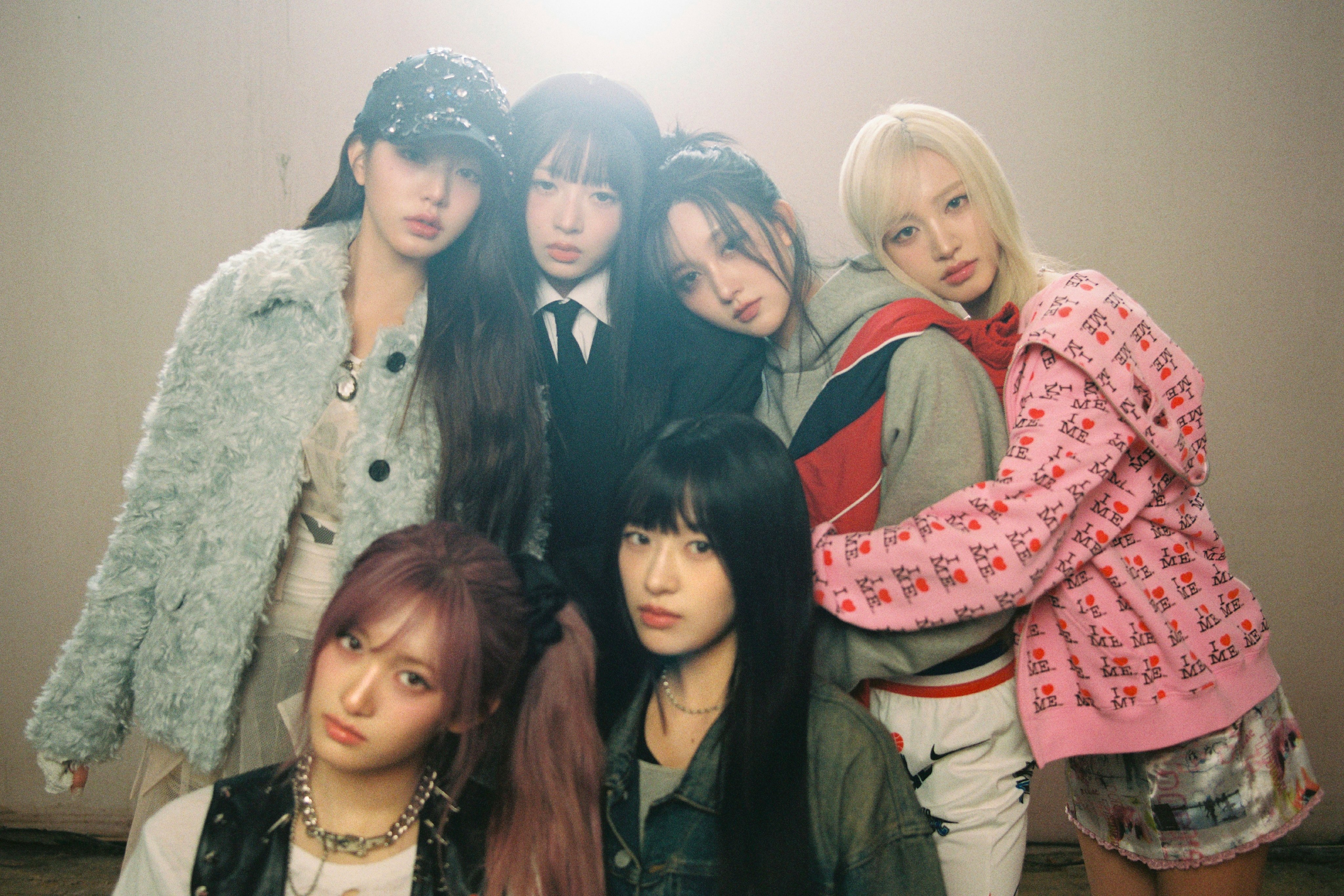 Seis jovens posam juntas em um ambiente interno com iluminação suave. Elas exibem estilos variados, combinando roupas casuais e modernas, com tons pastéis e urbanos, transmitindo uma atmosfera introspectiva e artística.